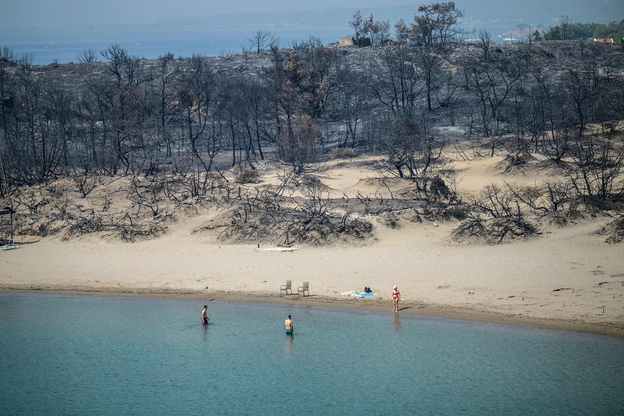 Een strand op het Griekse eiland Rhodos met op de achtergrond de door brand verwoeste bossen. Duizenden mensen zijn al van het eiland geëvacueerd.