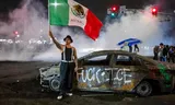 Een demonstrant zwaait zaterdag met een Mexicaanse vlag bij een uitgebrand autowrak vol graffiti gericht tegen de federale immigratiepolitie. Foto Ringo Chiu/AFP)  
